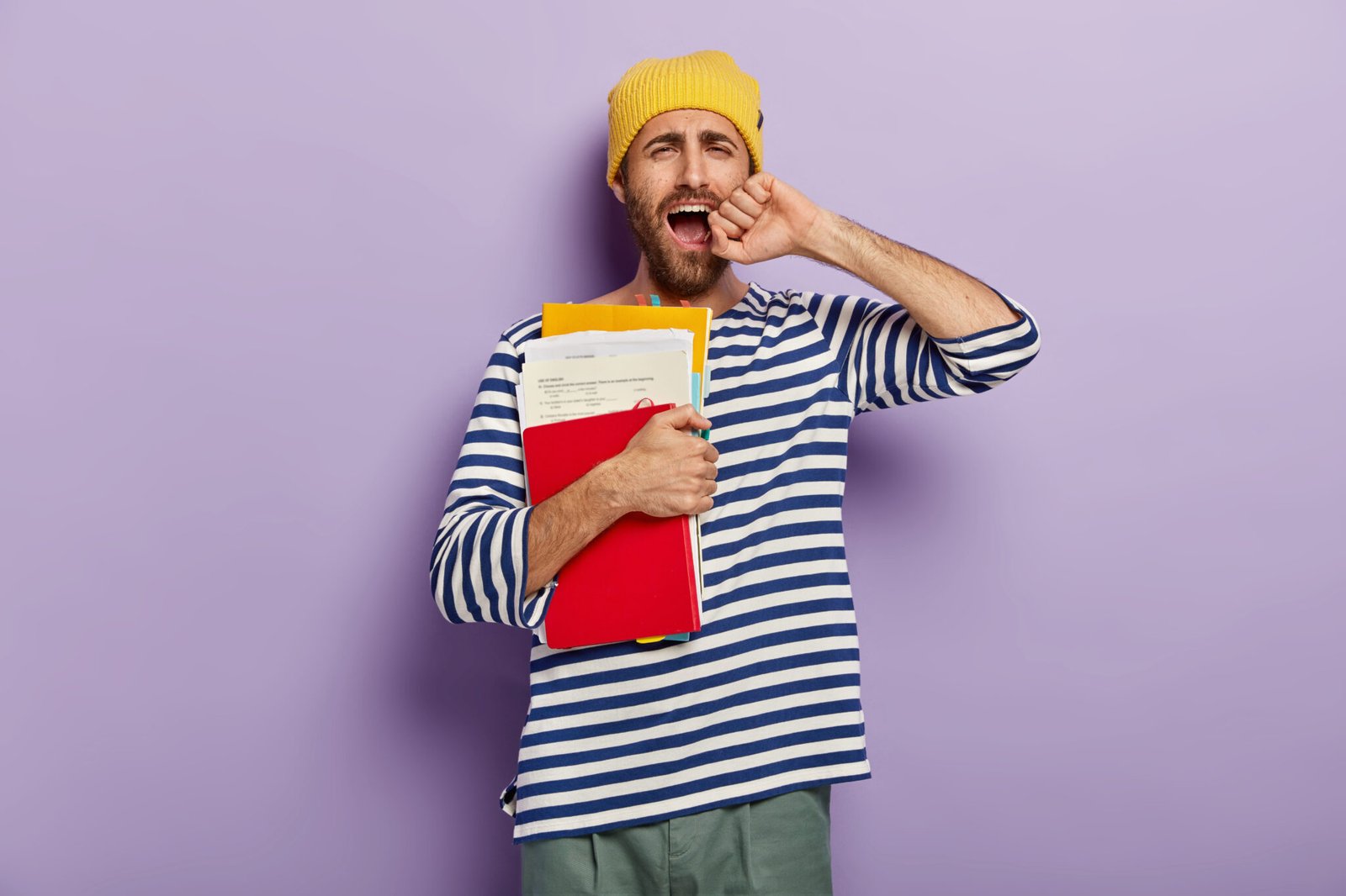 Photo of dissatisfied unshaven young man yawns from tiredness, being sleepy and exhausted, keeps hand near opened mouth, holds papers with notepad and textbook, isolated over purple background
